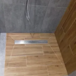 Wykończona kabina prysznicowa z odpływem liniowym, wyłożona płytkami imitującymi drewno na podłodze i ścianie, z szarymi płytkami na górnej części ściany.