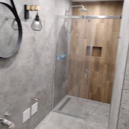 kajbud usługi remontowo-budowlane - Nowoczesna kabina prysznicowa z przesuwnymi drzwiami, wykończona płytkami imitującymi drewno i szarą mozaiką. Widoczny okrągły, czarny lustro i designerska lampa.