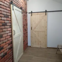 Dwie drewniane, przesuwne drzwi stodołowe z czarnymi okuciami. Jedne drzwi na tle ceglanej ściany, drugie na tle białej ściany. Jasna podłoga.