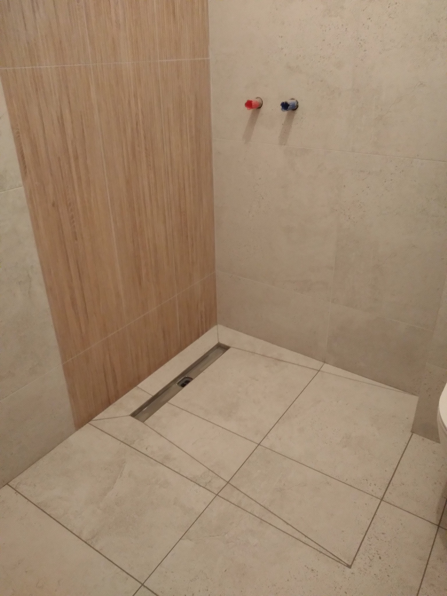 Nowoczesna kabina prysznicowa bez brodzika z odpływem liniowym i płytkami w odcieniach beżu i brązu. Dwa przyłącza wody na ścianie.