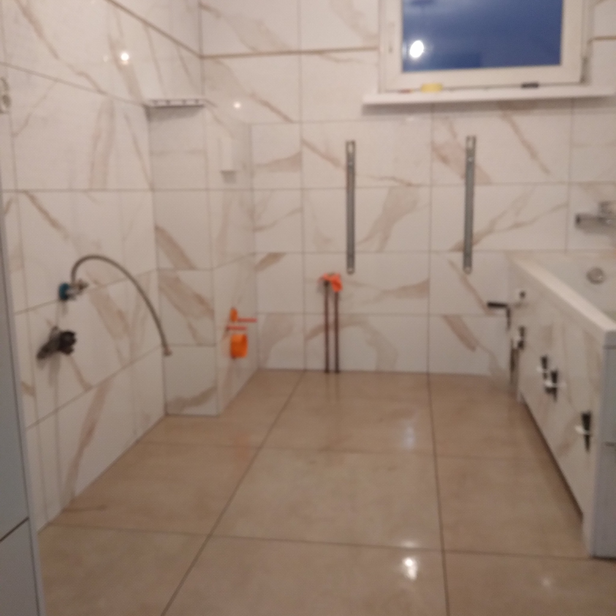 Wnętrze łazienki w trakcie prac: widoczne rury, instalacja wodna, białe marmurowe płytki na ścianach i beżowe na podłodze. Okno w tle.