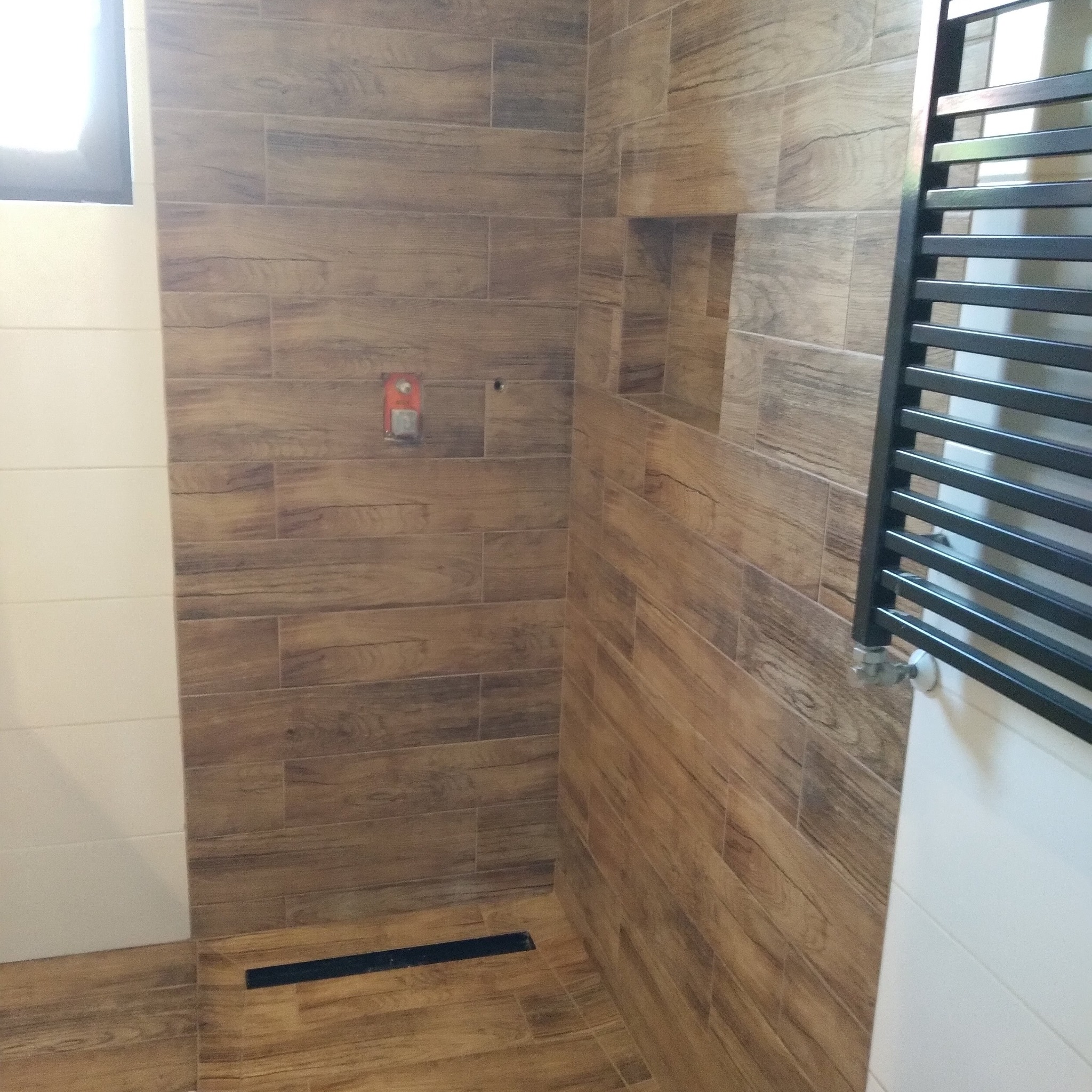 Wykończona łazienka z drewnopodobnymi płytkami na ścianach i podłodze. Widoczny grzejnik drabinkowy i wnęka w ścianie. Minimalistyczny odpływ liniowy.