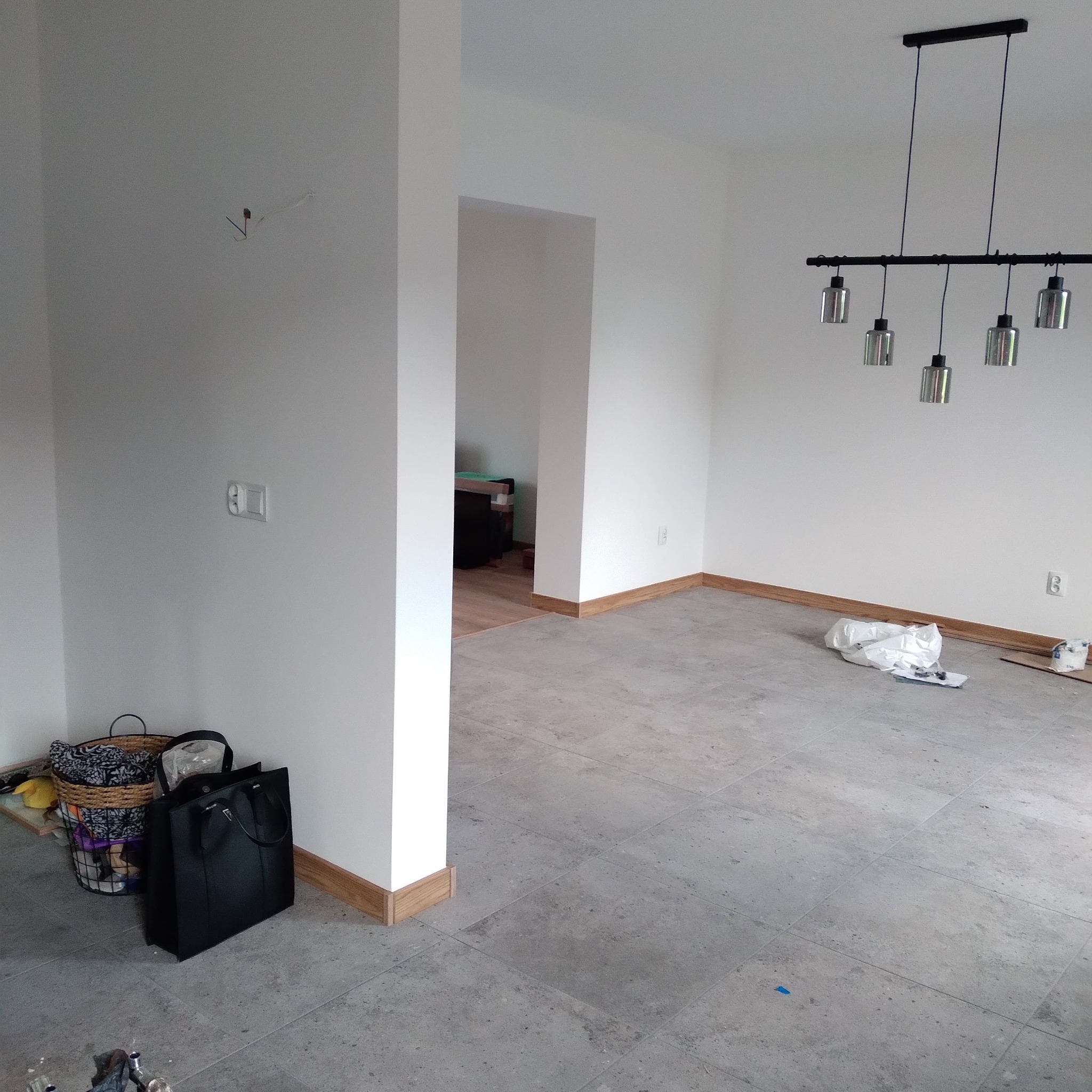 Jasne, prawie puste pomieszczenie z szarą podłogą z płytek, białe ściany z drewnianymi listwami przypodłogowymi i nowoczesną lampą sufitową.