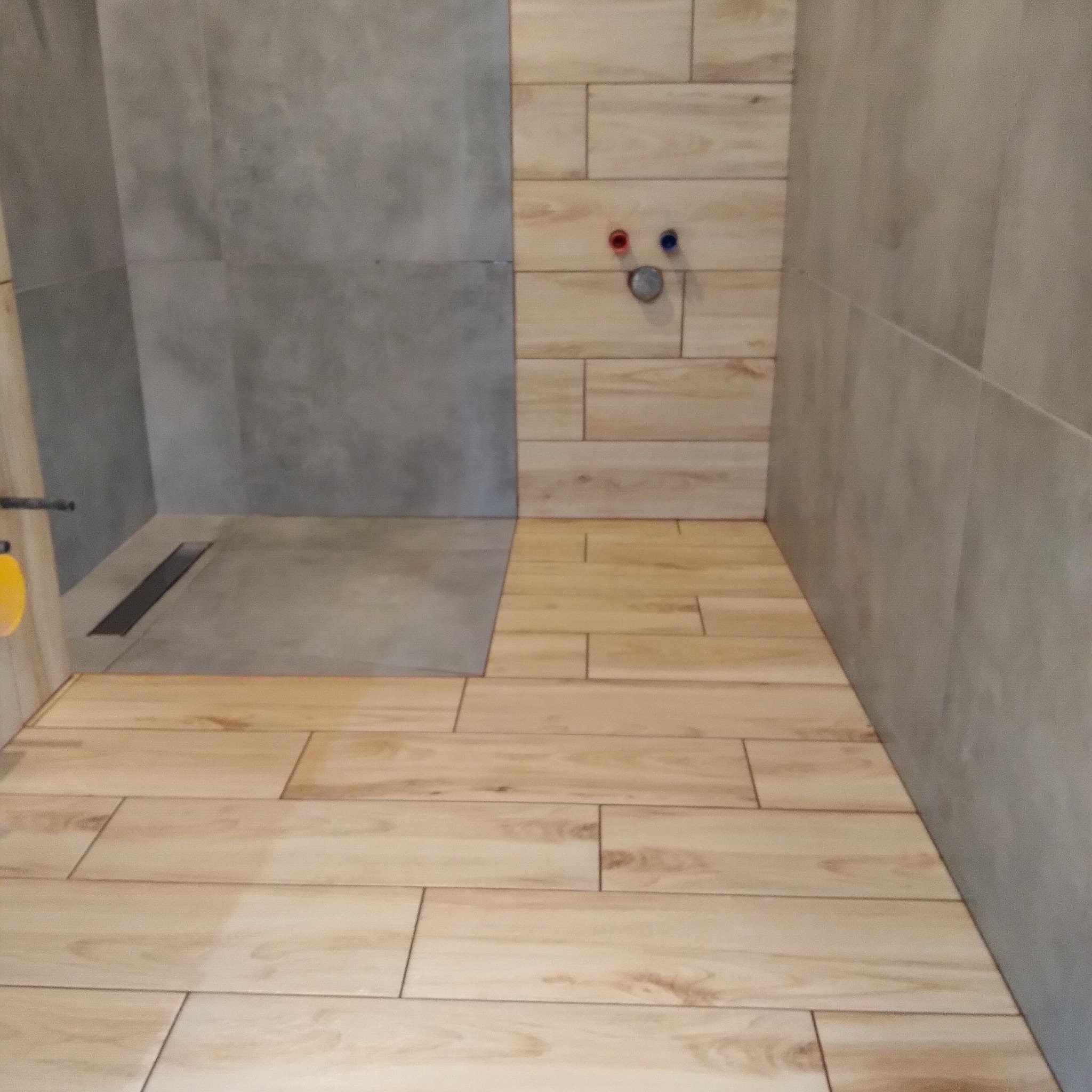 Ukończona łazienka z drewnopodobnymi płytkami podłogowymi i szarą strefą prysznicową z odpływem liniowym. Widoczne przyłącza wodne na ścianie.