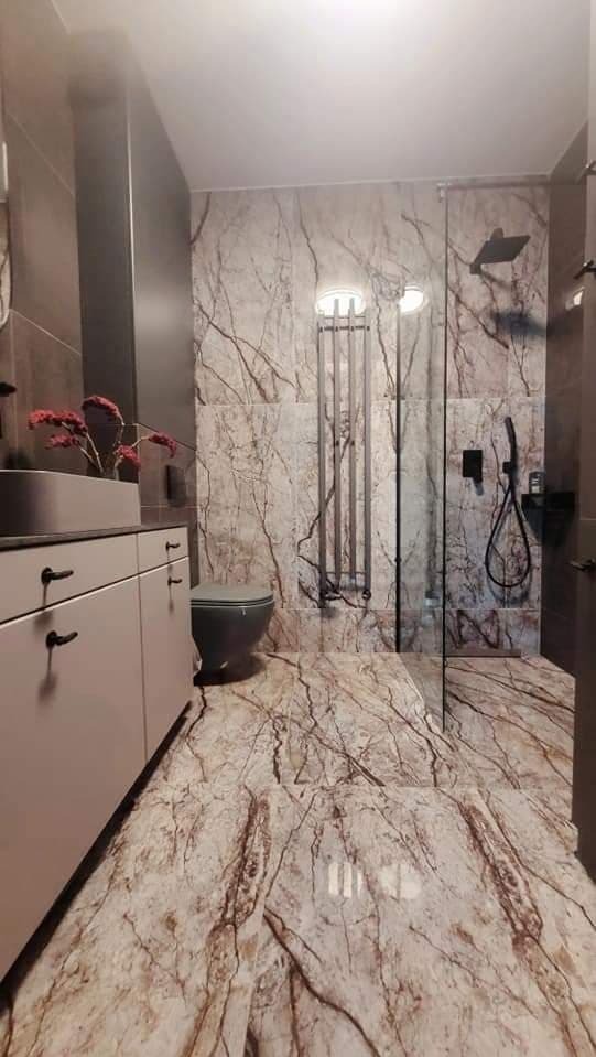 Nowoczesna łazienka z marmurową podłogą i ścianami, szklaną kabiną prysznicową oraz grafitową toaletą i umywalką. Minimalistyczny design i eleganckie wykończenie.