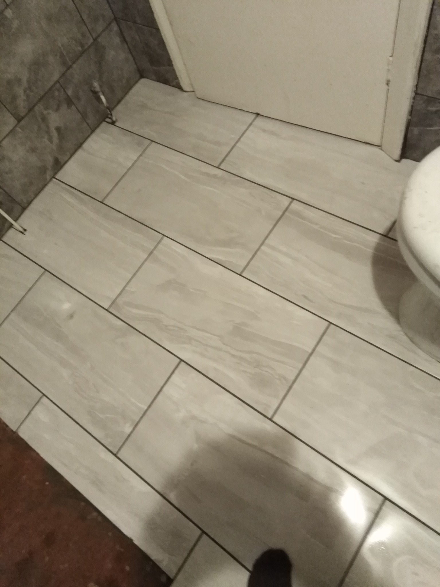 Ułożona pod kątem jasnoszara podłoga z płytek imitujących drewno w łazience, widoczny fragment toalety i szare płytki na ścianie.