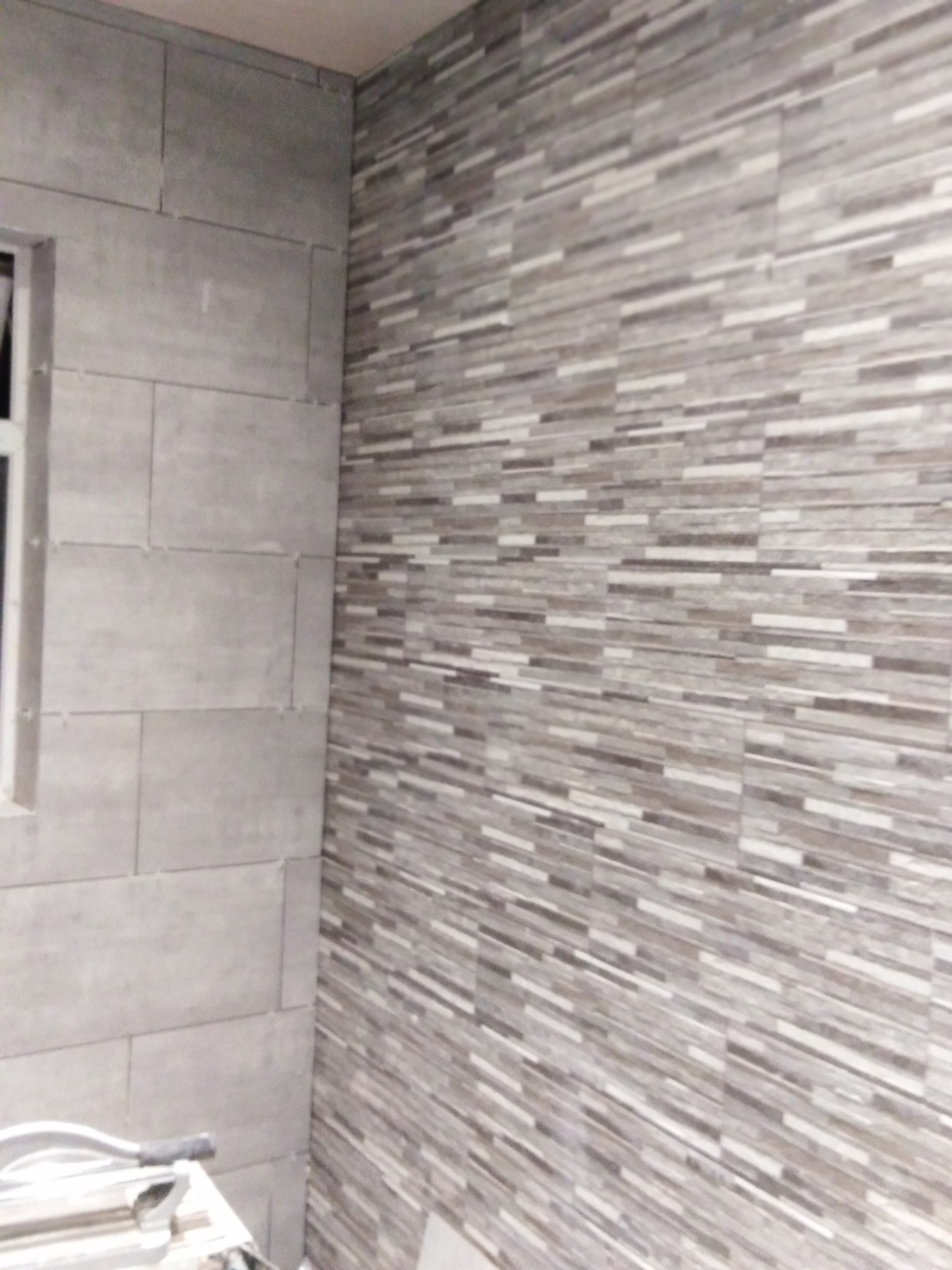 Ściana łazienki wyłożona szarymi, poziomymi płytkami imitującymi kamień, z widocznym fragmentem okna i narzędzia do cięcia płytek.