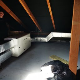 KOVALINSTAL JACEK KOWALIK - Instalacja rekuperacji na poddaszu: widoczny centralny moduł, srebrne rury wentylacyjne z folii, drewniane elementy konstrukcyjne dachu, osoba w tle. Słabe oświetlenie.