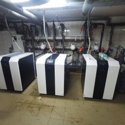 KOVALINSTAL JACEK KOWALIK - Trzy nowoczesne pompy ciepła w kotłowni z rurami i armaturą. Biało-czarne obudowy, tło z płytek. Widok na instalację po montażu.