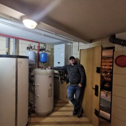 KOVALINSTAL JACEK KOWALIK - Instalacja grzewcza w kotłowni z zasobnikami ciepłej wody i piecem gazowym. Mężczyzna opiera się o zasobnik, w tle widoczne rury i instalacje.