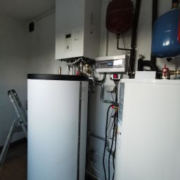 KOVALINSTAL JACEK KOWALIK - Biały, wiszący piec kondensacyjny Panasonic Aquarea z zbiornikiem na wodę, instalacją hydrauliczną i elektryczną w kotłowni. Widoczna drabina.