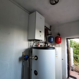 KOVALINSTAL JACEK KOWALIK - Biały piec grzewczy i zbiornik na wodę w kotłowni, z widocznymi rurami, manometrami i elementami instalacji. W tle widoczne dodatkowe zbiorniki i otwarte drzwi.