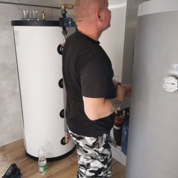 KOVALINSTAL JACEK KOWALIK - Instalator przy dwóch stojących, białych i srebrnych, zbiornikach na wodę w pomieszczeniu z brązowymi płytkami podłogowymi. Widoczny manometr.