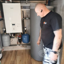 KOVALINSTAL JACEK KOWALIK - Biały piec Panasonic z szarym zbiornikiem i niebieskim naczyniem przeponowym na tle beżowych płytek. Instalator w czarnej koszulce obok zbiornika.