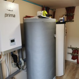 KOVALINSTAL JACEK KOWALIK - Kotłownia z szarym zasobnikiem CWU, białym kotłem Galmet Prima i rozdzielaczami CO z czerwonymi rurami. Widoczne rury i instalacje pod sufitem.