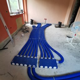 KOVALINSTAL JACEK KOWALIK - Niebieskie rury rekuperacji rozłożone na podłodze w trakcie montażu, widoczne skrzynki rozdzielcze i fragment okna. Pomieszczenie w trakcie remontu.