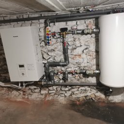 KOVALINSTAL JACEK KOWALIK - Biały bojler i piec Panasonic na tle ściany z cegły, szare rury i instalacja. Widoczny podłączony przedłużacz z kolorowymi przyciskami.