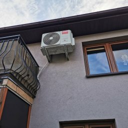 KOVALINSTAL JACEK KOWALIK - Biały klimatyzator VIVAX zamontowany na szarej elewacji budynku obok okna z brązową ramą i balkonu z czarną balustradą. Widok z dołu na błękitne niebo.