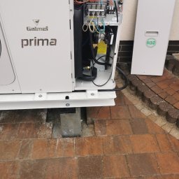 KOVALINSTAL JACEK KOWALIK - Otwarta obudowa pompy ciepła Galmet Prima z widocznymi elementami wewnętrznymi i elektroniką. Na obudowie widoczny napis R32. Pompa stoi na kostce brukowej.