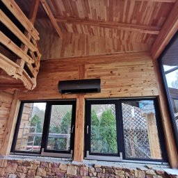 KOVALINSTAL JACEK KOWALIK - Klimatyzator na drewnianej ścianie w pomieszczeniu z oknami. Widoczne elementy drewnianej konstrukcji i kamienna podmurówka. Na zewnątrz zieleń.