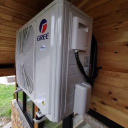 KOVALINSTAL JACEK KOWALIK - Klimatyzator GREE zamontowany na zewnątrz budynku z drewnianym wykończeniem, widoczny z boku, z fragmentem zieleni w tle.