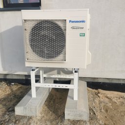 KOVALINSTAL JACEK KOWALIK - Zewnętrzna jednostka pompy ciepła Panasonic Inverter, zamontowana na białym stelażu na betonowym fundamencie przy ścianie budynku. Widoczne oznaczenie R32.