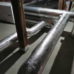 KOVALINSTAL JACEK KOWALIK - Szczegółowe ujęcie izolowanych, aluminiowych rur wentylacyjnych w piwnicy, z widocznym urządzeniem rekuperacyjnym w tle i drewnianym wspornikiem.