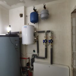 KOVALINSTAL JACEK KOWALIK - Biały zasobnik ciepłej wody użytkowej z szarym i niebieskim zbiornikiem przeponowym oraz orurowaniem w surowym pomieszczeniu z niedokończonymi ścianami.