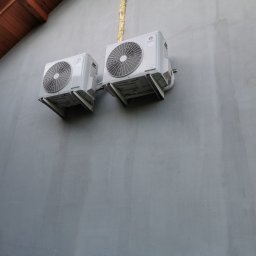 KOVALINSTAL JACEK KOWALIK - Dwa białe klimatyzatory GREE zamontowane na szarej ścianie budynku z brązowym fragmentem dachu w rogu. Widoczna instalacja odprowadzająca skropliny.