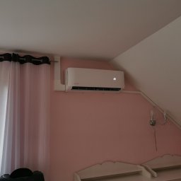KOVALINSTAL JACEK KOWALIK - Klimatyzator Rotenso z wyświetlaczem 23°C zamontowany na różowej ścianie pod skosem, widoczna lampka nocna i zasłony. Widok na poddasze.