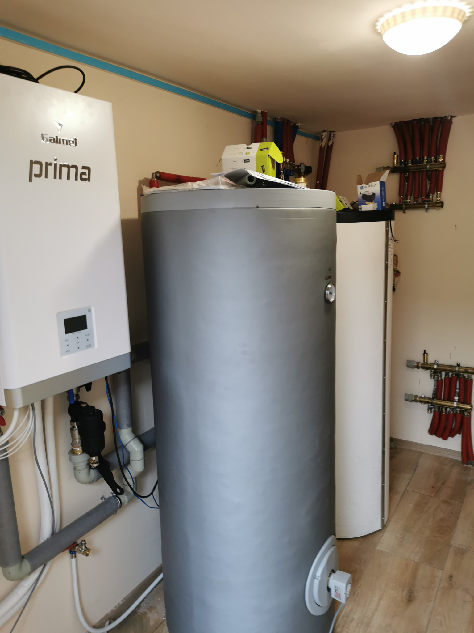 Kotłownia z szarym zasobnikiem CWU, białym kotłem Galmet Prima i rozdzielaczami CO z czerwonymi rurami. Widoczne rury i instalacje pod sufitem.