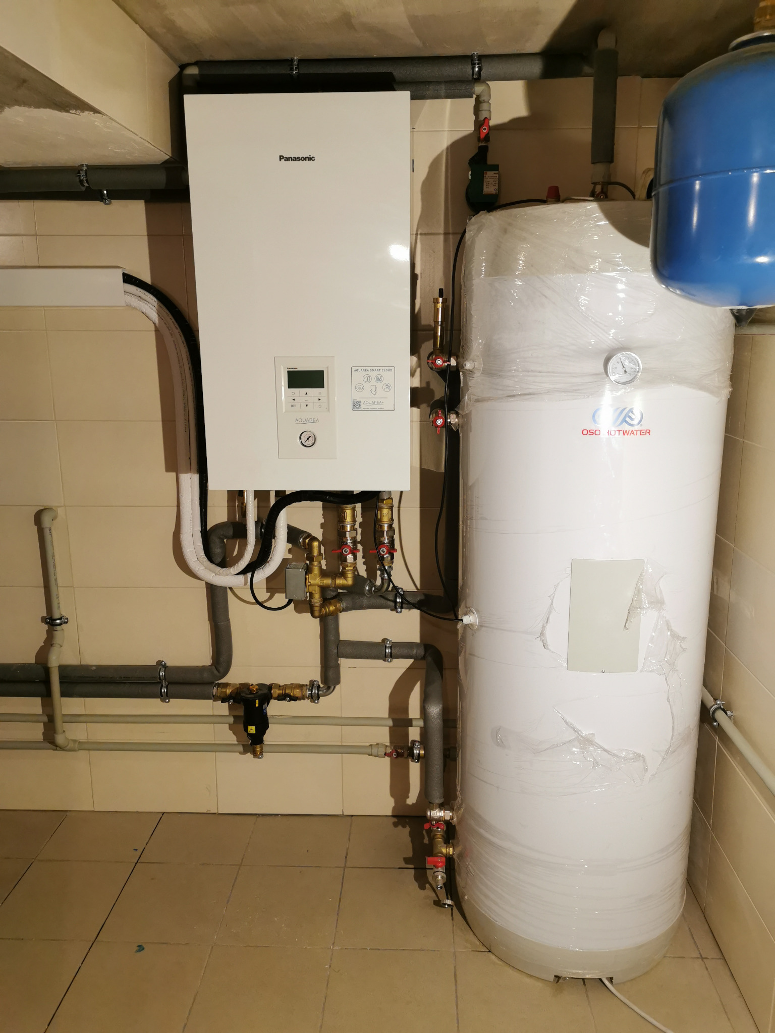Biały, wiszący piec Panasonic Aquarea Smart Cloud z podłączonymi rurami i zbiornikiem OSO Hotwater w pomieszczeniu z kafelkami. Widoczne elementy instalacji.