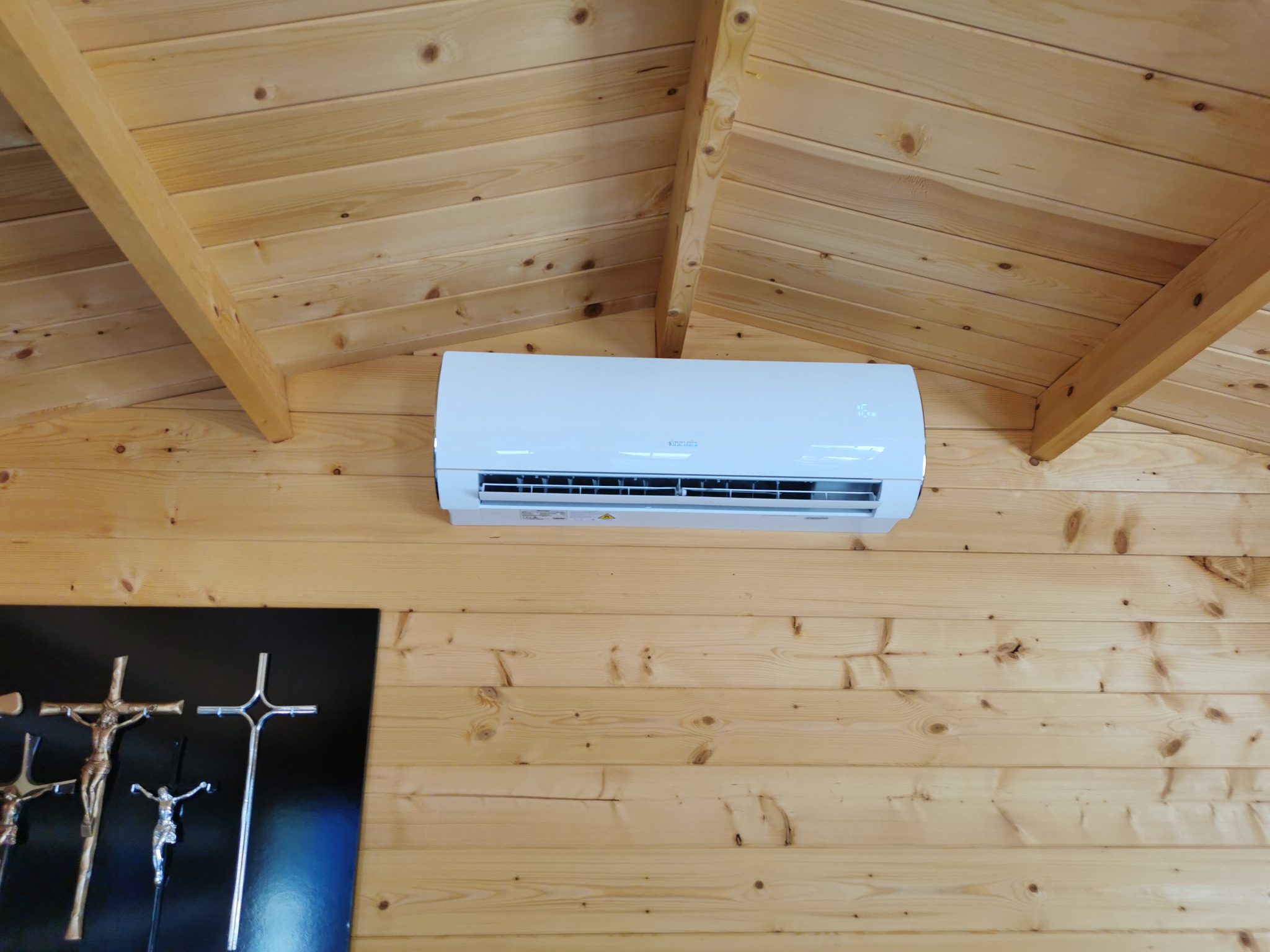 Biały klimatyzator Sinclair zamontowany na drewnianej ścianie pod skośnym, drewnianym sufitem, z elementami dekoracyjnymi po lewej stronie.
