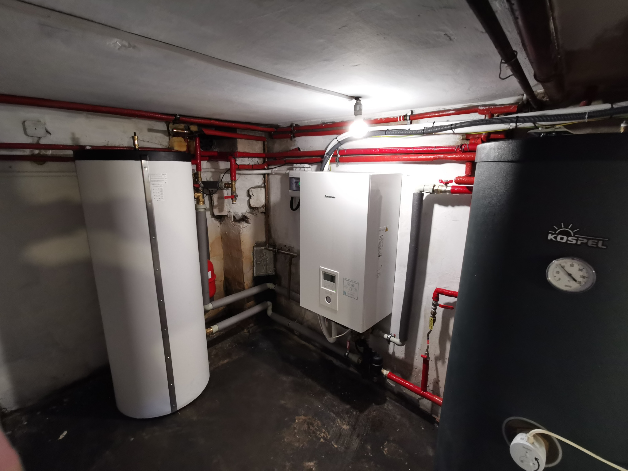 Kotłownia: biały, wiszący piec Panasonic, obok biały i czarny zbiornik Kospel. Czerwone rury na ścianach i suficie. Widoczne przyłącza i instalacje.