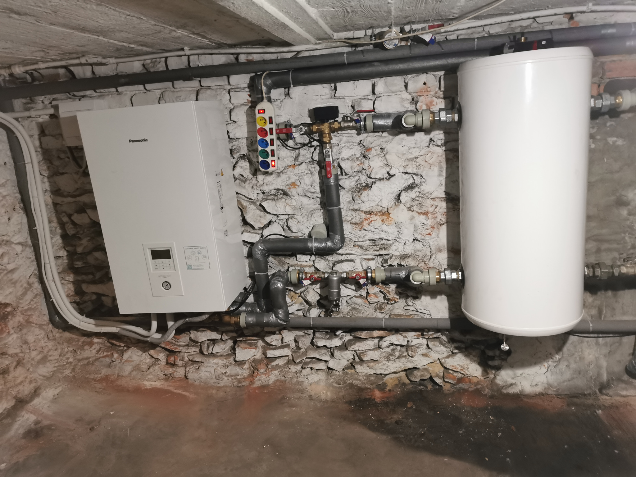 Biały bojler i piec Panasonic na tle ściany z cegły, szare rury i instalacja. Widoczny podłączony przedłużacz z kolorowymi przyciskami.
