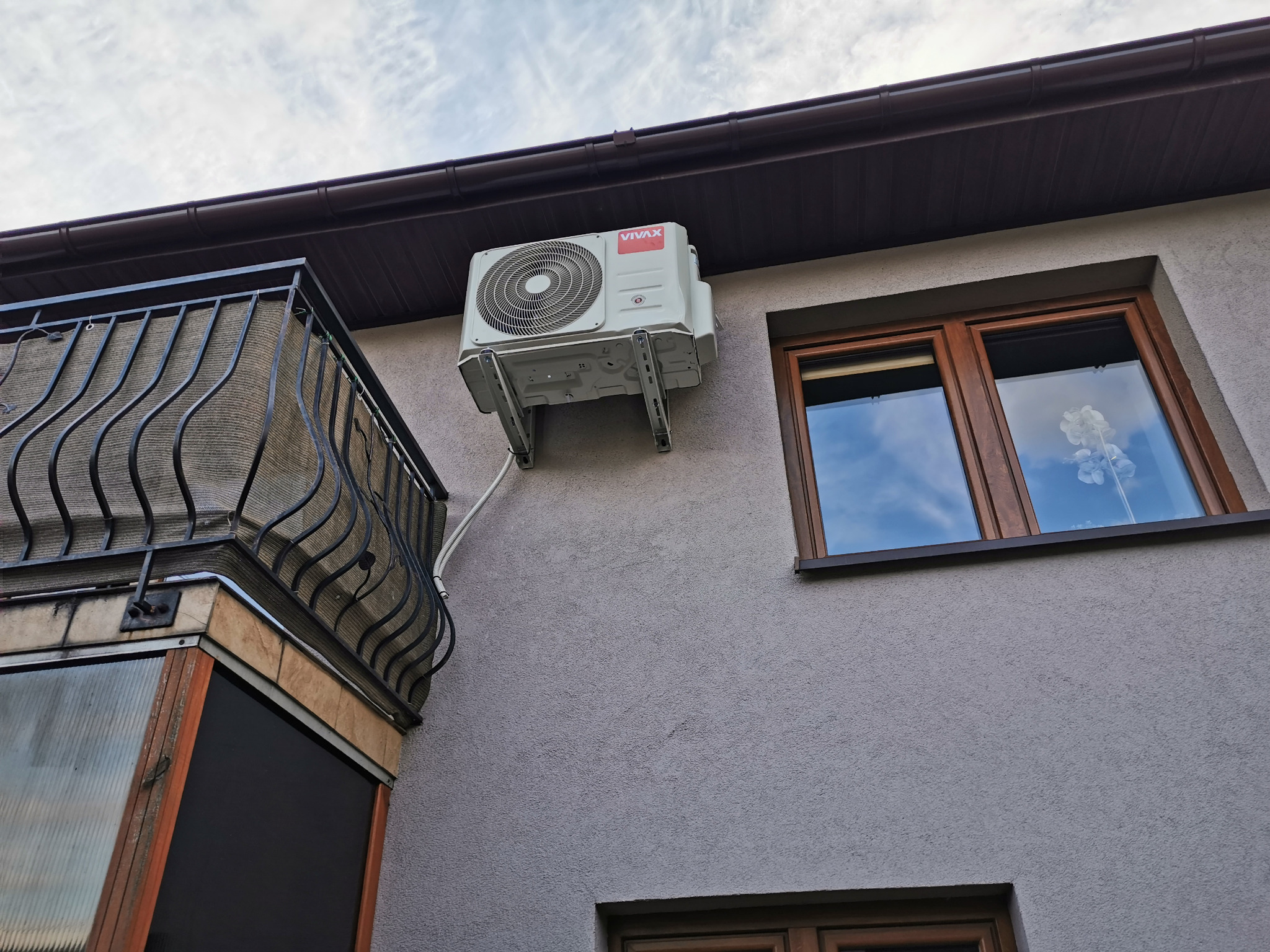 Biały klimatyzator VIVAX zamontowany na szarej elewacji budynku obok okna z brązową ramą i balkonu z czarną balustradą. Widok z dołu na błękitne niebo.