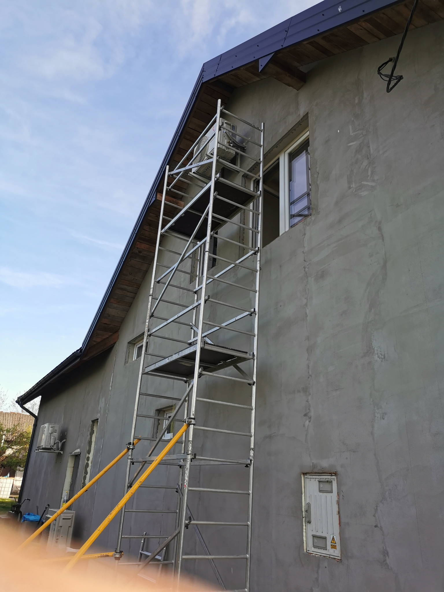 Dom z szarą elewacją w trakcie wykończenia, widoczne rusztowanie, okno i klimatyzator na ścianie. Na górze kabel elektryczny.