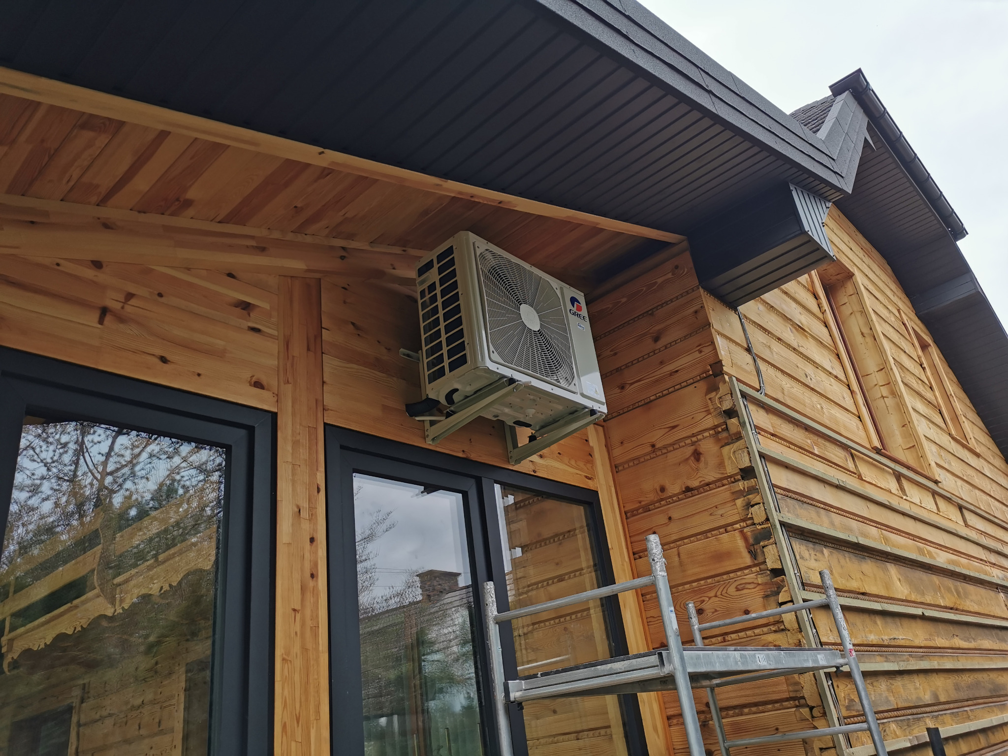 Montaż klimatyzacji zewnętrznej na drewnianej elewacji domu z widoczną drabiną, wykonany pod kątem, prezentujący detale instalacji i otoczenia.
