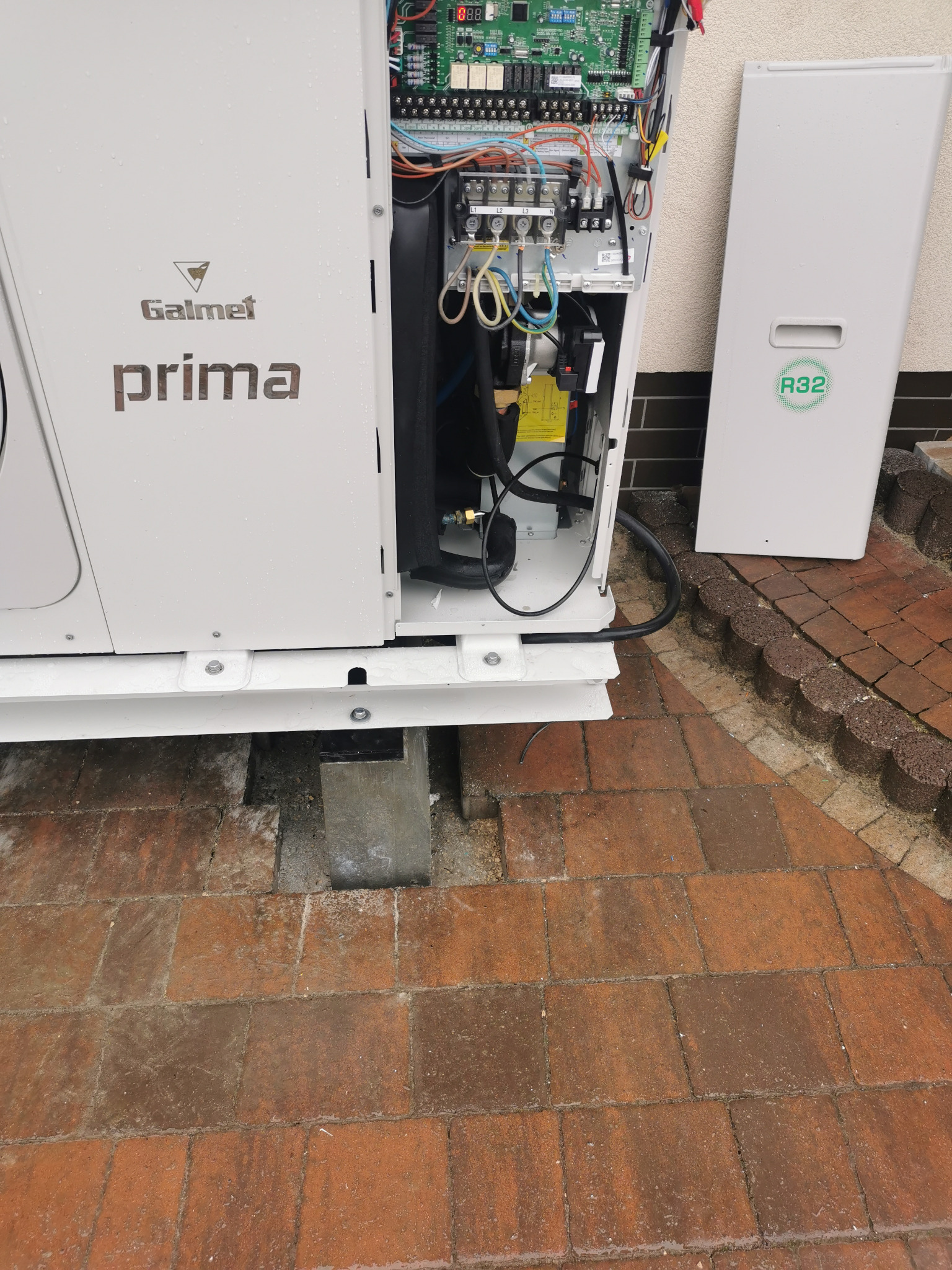 Otwarta obudowa pompy ciepła Galmet Prima z widocznymi elementami wewnętrznymi i elektroniką. Na obudowie widoczny napis R32. Pompa stoi na kostce brukowej.