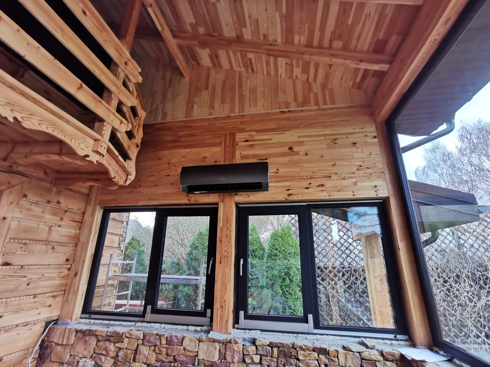 Klimatyzator na drewnianej ścianie w pomieszczeniu z oknami. Widoczne elementy drewnianej konstrukcji i kamienna podmurówka. Na zewnątrz zieleń.