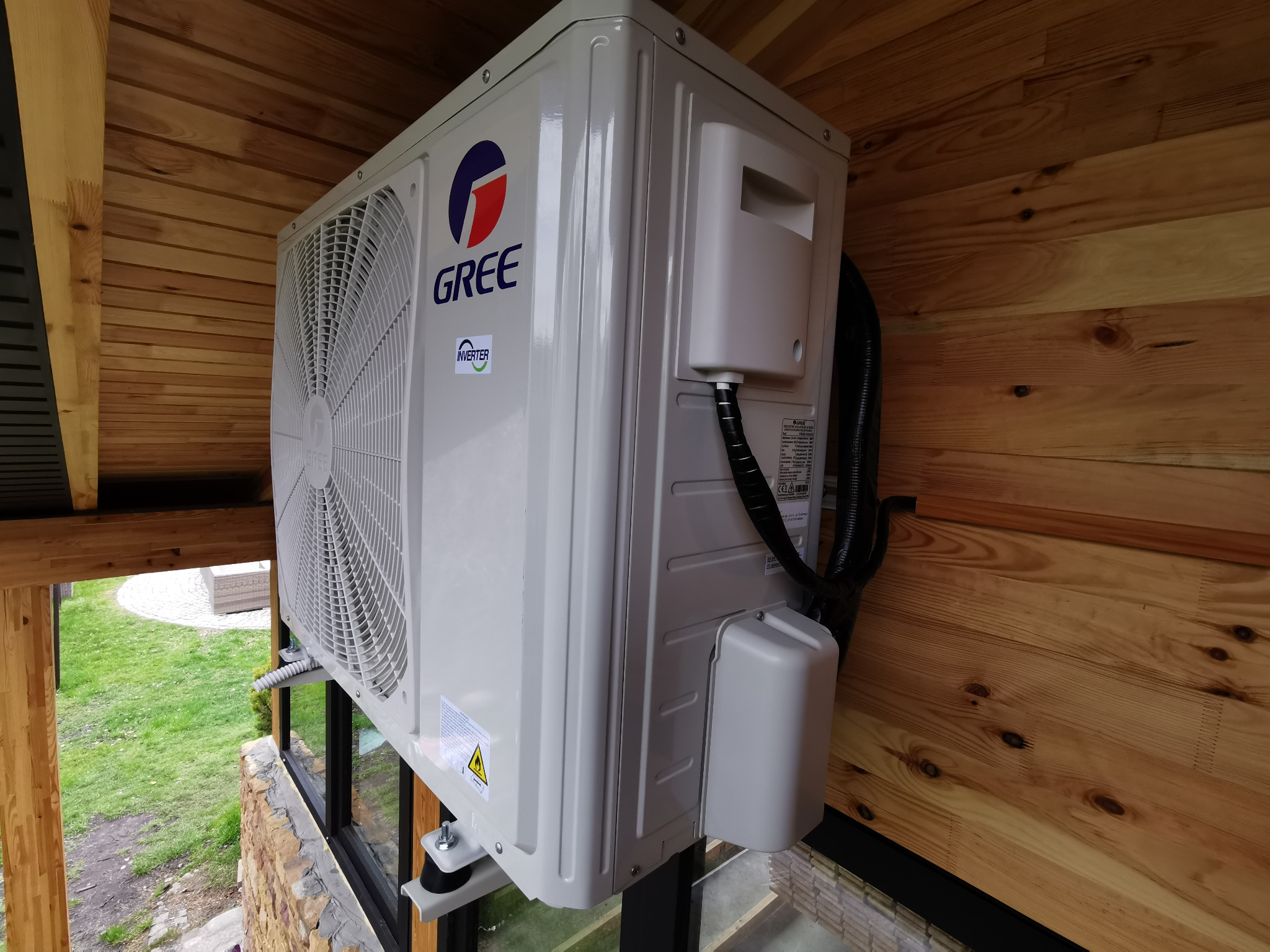 Klimatyzator GREE zamontowany na zewnątrz budynku z drewnianym wykończeniem, widoczny z boku, z fragmentem zieleni w tle.
