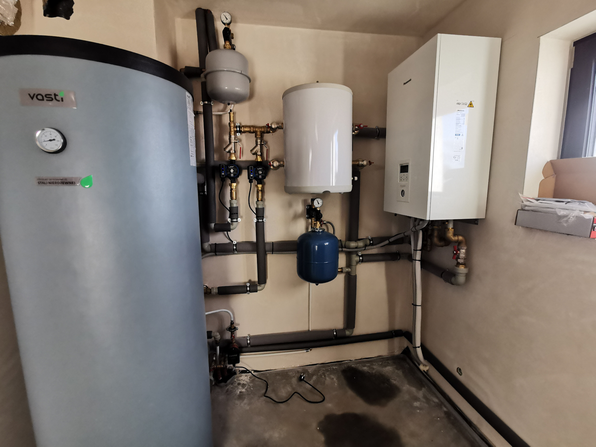 Zestaw urządzeń grzewczych: zbiornik Vasti, zasobnik wody, pompa ciepła Panasonic, rury z izolacją, manometry, zawory. Kompaktowa instalacja w pomieszczeniu technicznym.