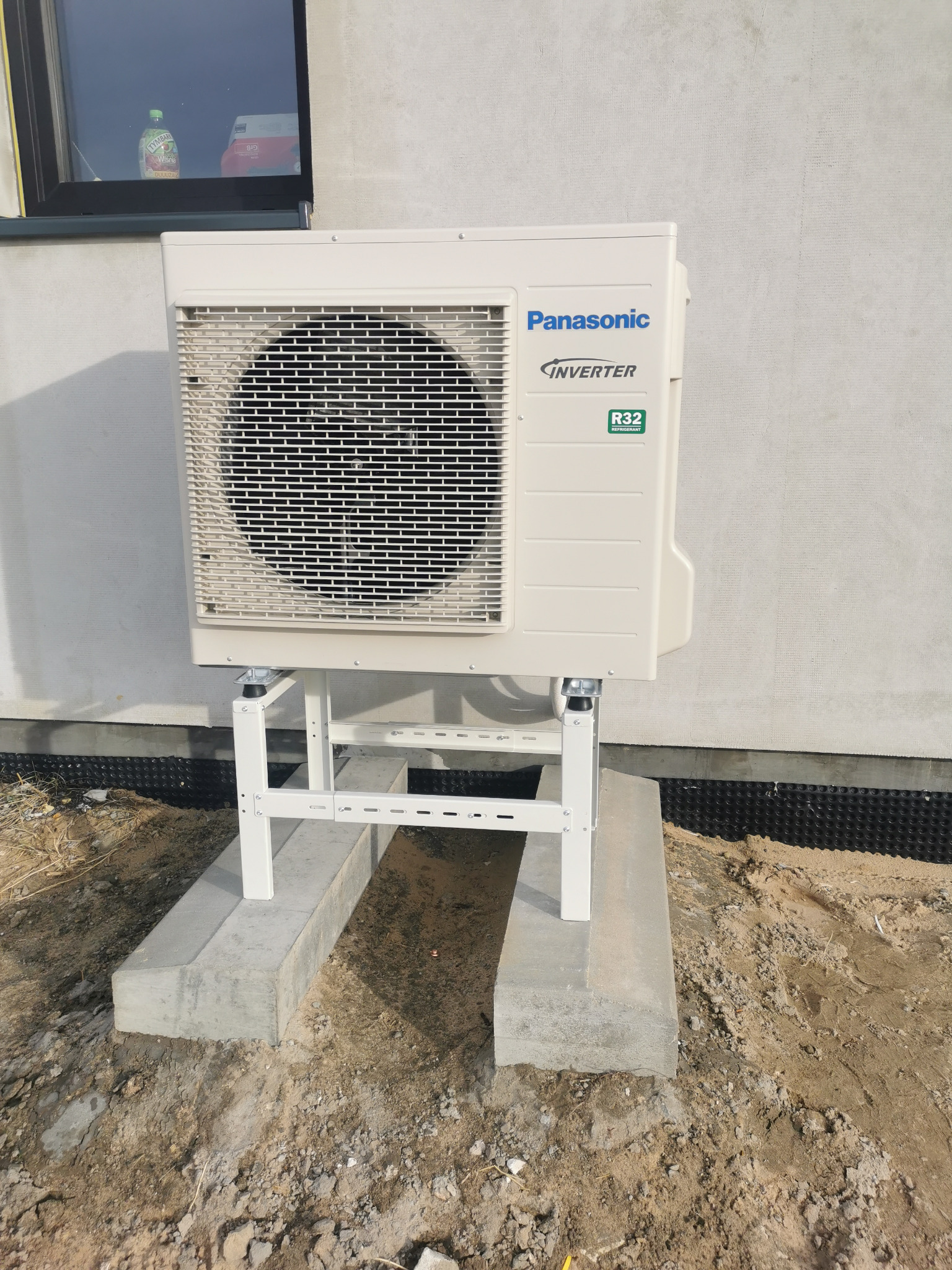 Zewnętrzna jednostka pompy ciepła Panasonic Inverter, zamontowana na białym stelażu na betonowym fundamencie przy ścianie budynku. Widoczne oznaczenie R32.