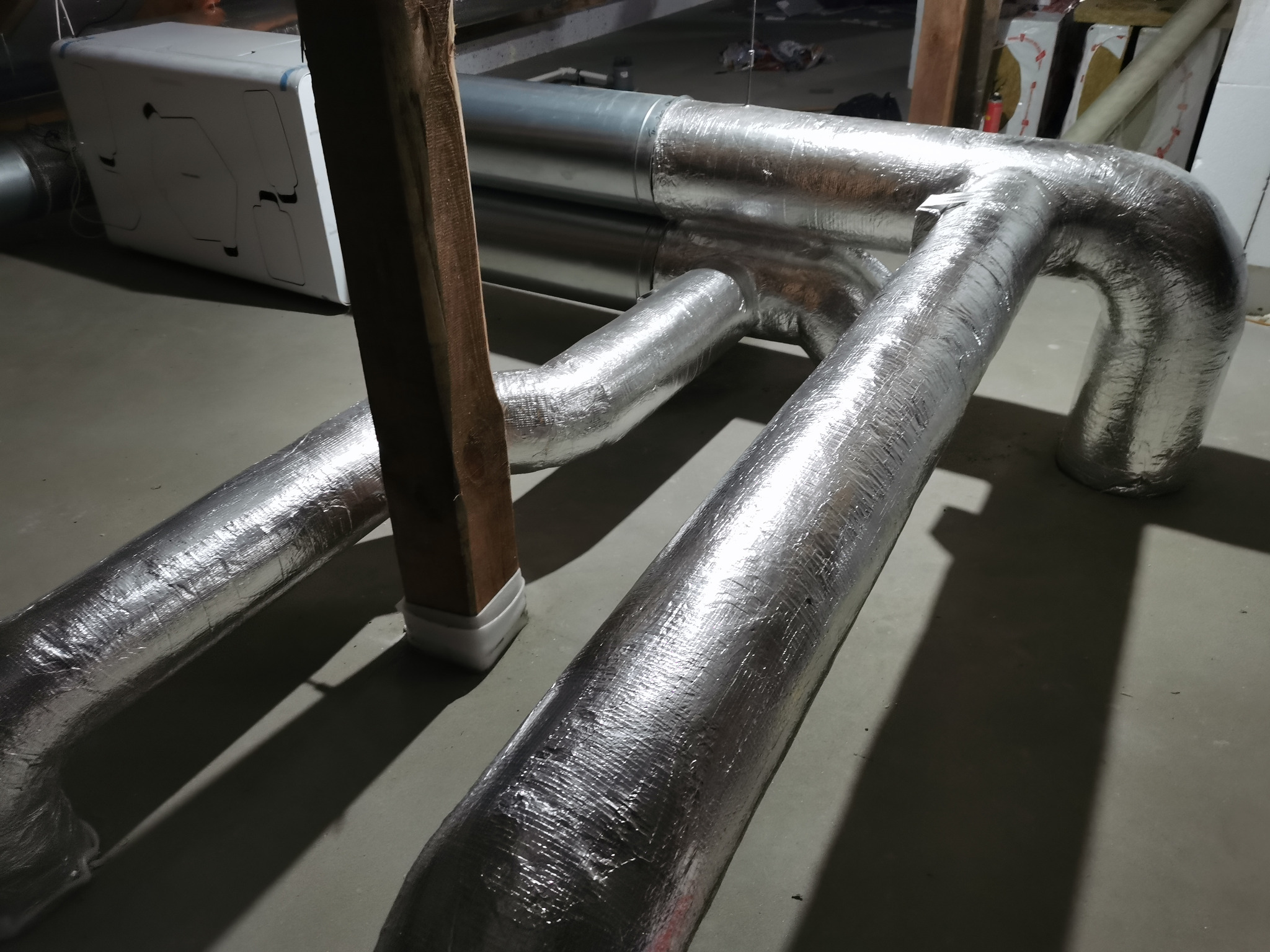 Szczegółowe ujęcie izolowanych, aluminiowych rur wentylacyjnych w piwnicy, z widocznym urządzeniem rekuperacyjnym w tle i drewnianym wspornikiem.