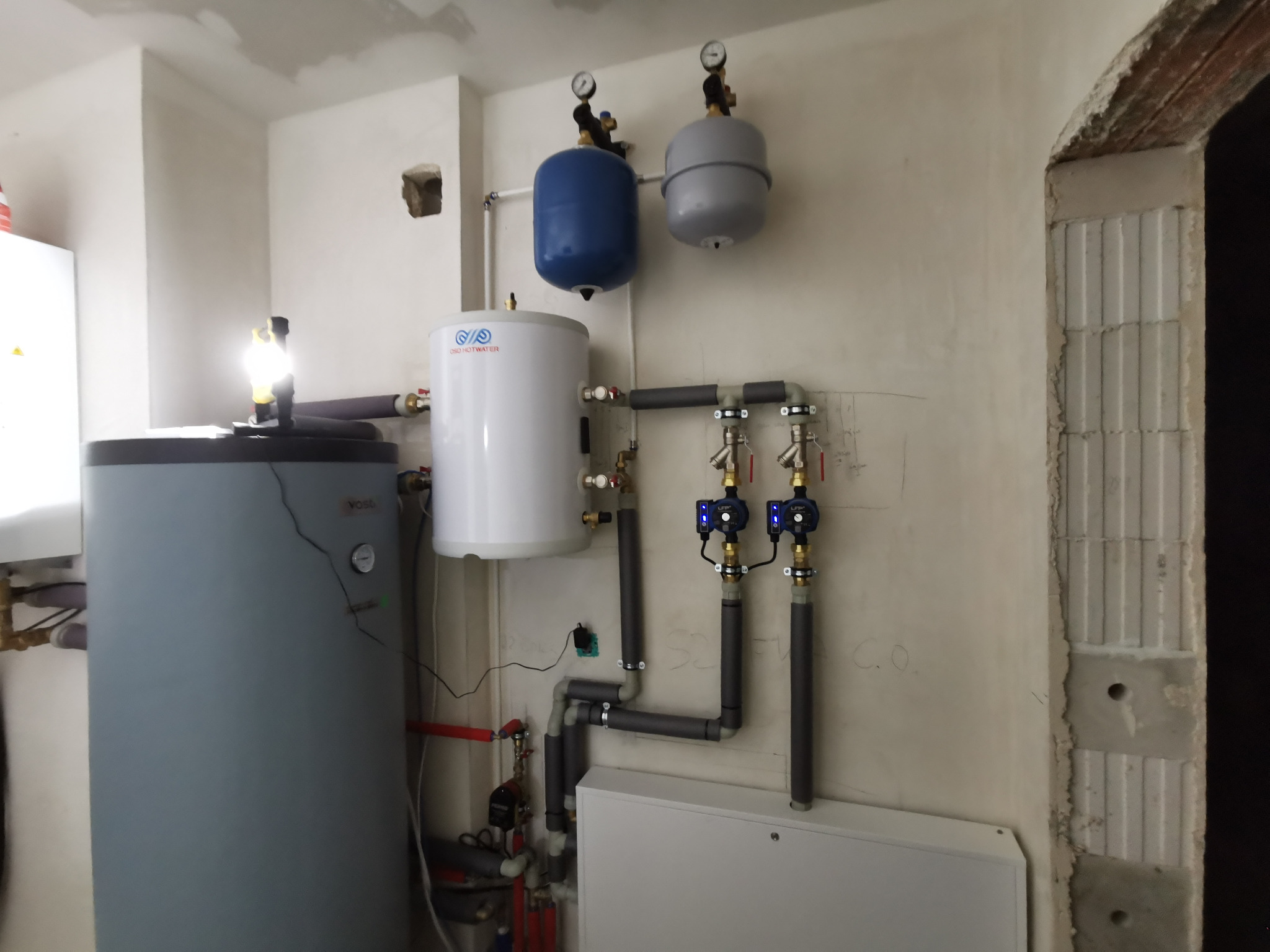 Biały zasobnik ciepłej wody użytkowej z szarym i niebieskim zbiornikiem przeponowym oraz orurowaniem w surowym pomieszczeniu z niedokończonymi ścianami.