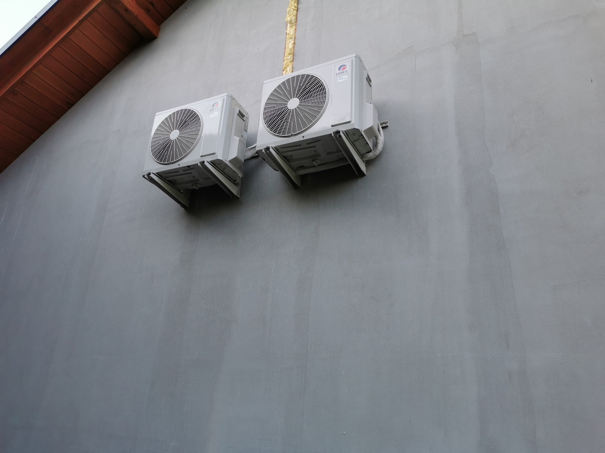Dwa białe klimatyzatory GREE zamontowane na szarej ścianie budynku z brązowym fragmentem dachu w rogu. Widoczna instalacja odprowadzająca skropliny.