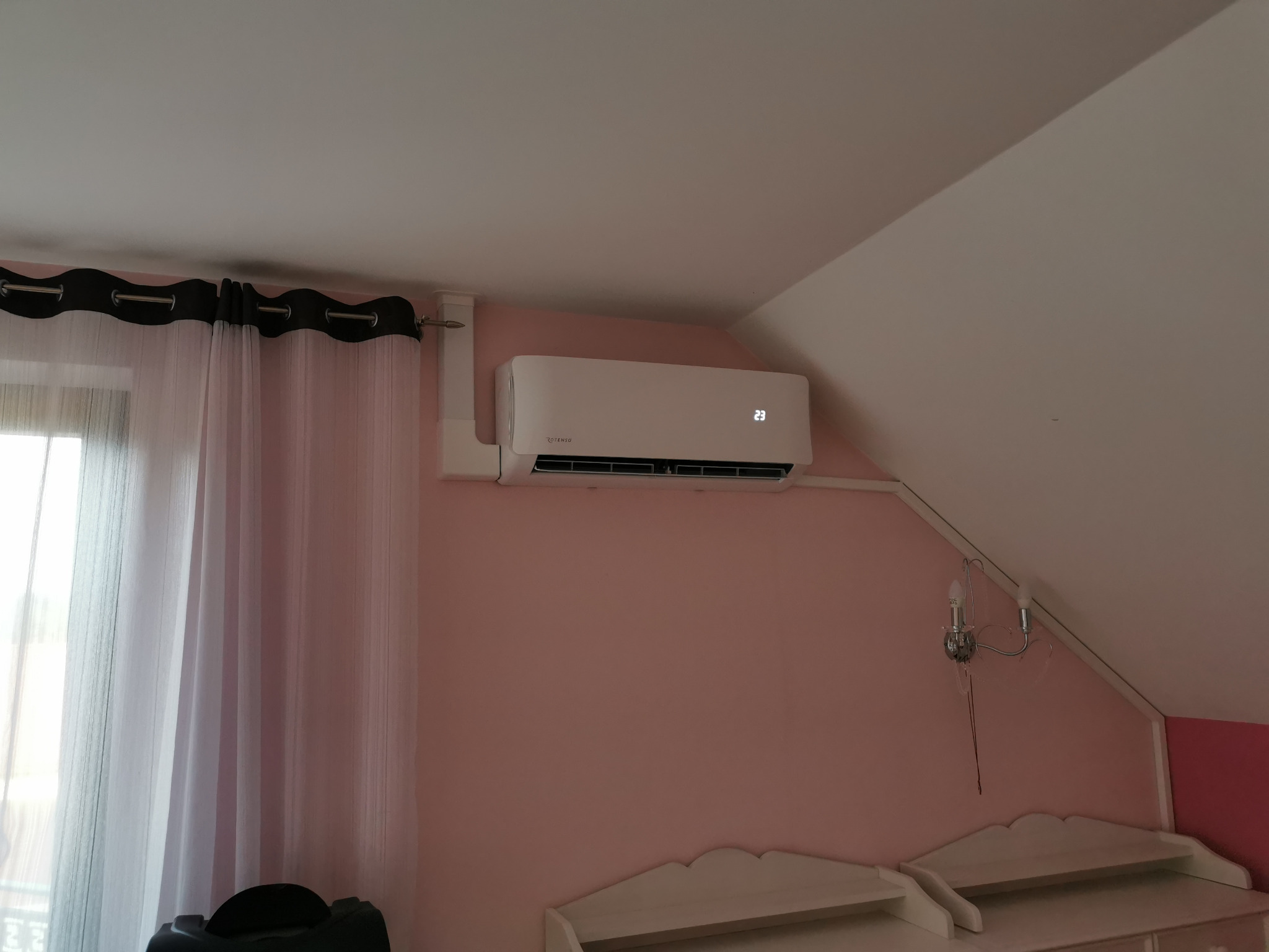 Klimatyzator Rotenso z wyświetlaczem 23°C zamontowany na różowej ścianie pod skosem, widoczna lampka nocna i zasłony. Widok na poddasze.