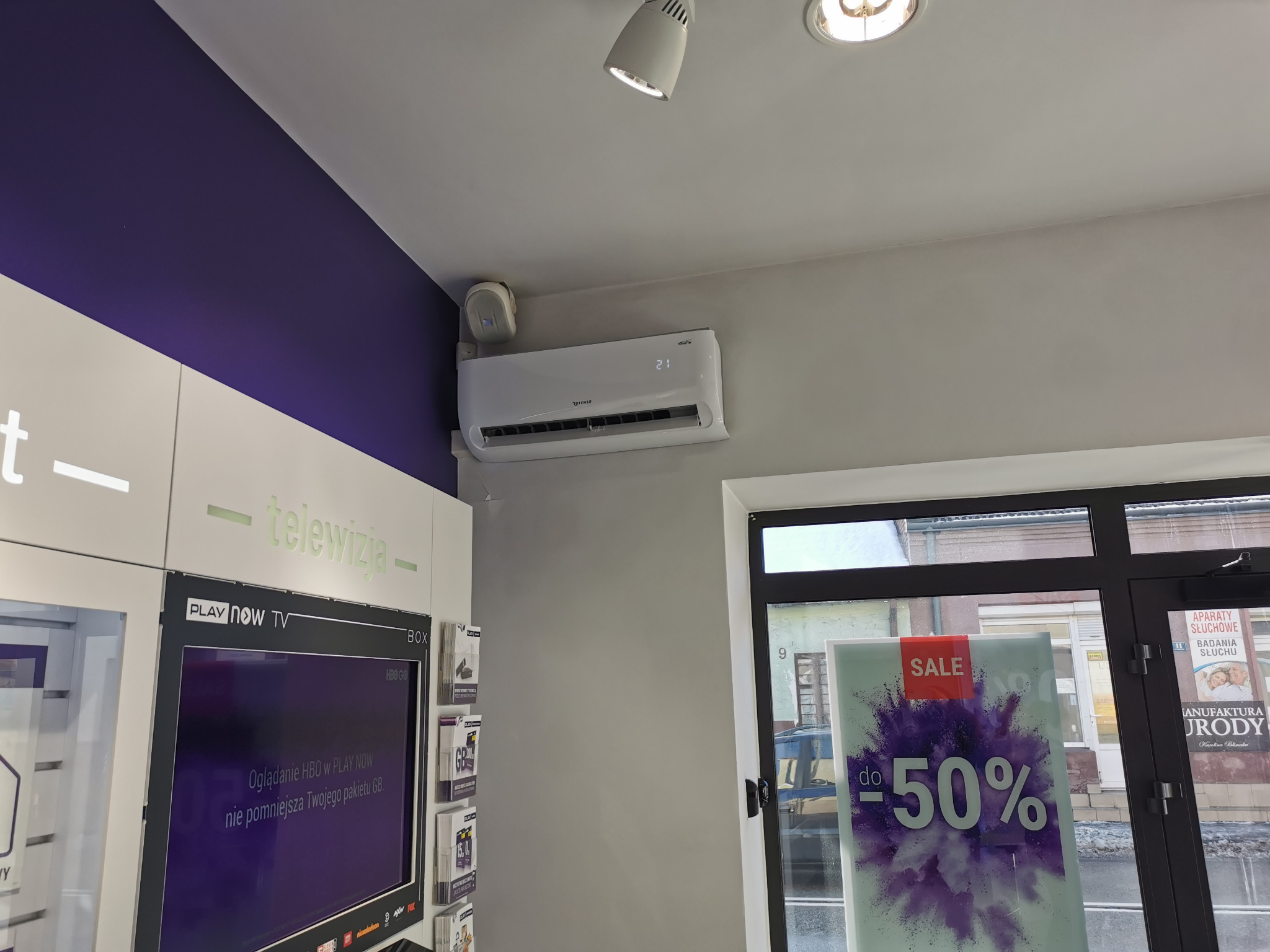 Klimatyzator Rotenso zamontowany na ścianie w pomieszczeniu z fioletowym akcentem kolorystycznym. Widoczny ekran telewizora i szyba z napisem 'SALE -50%'.