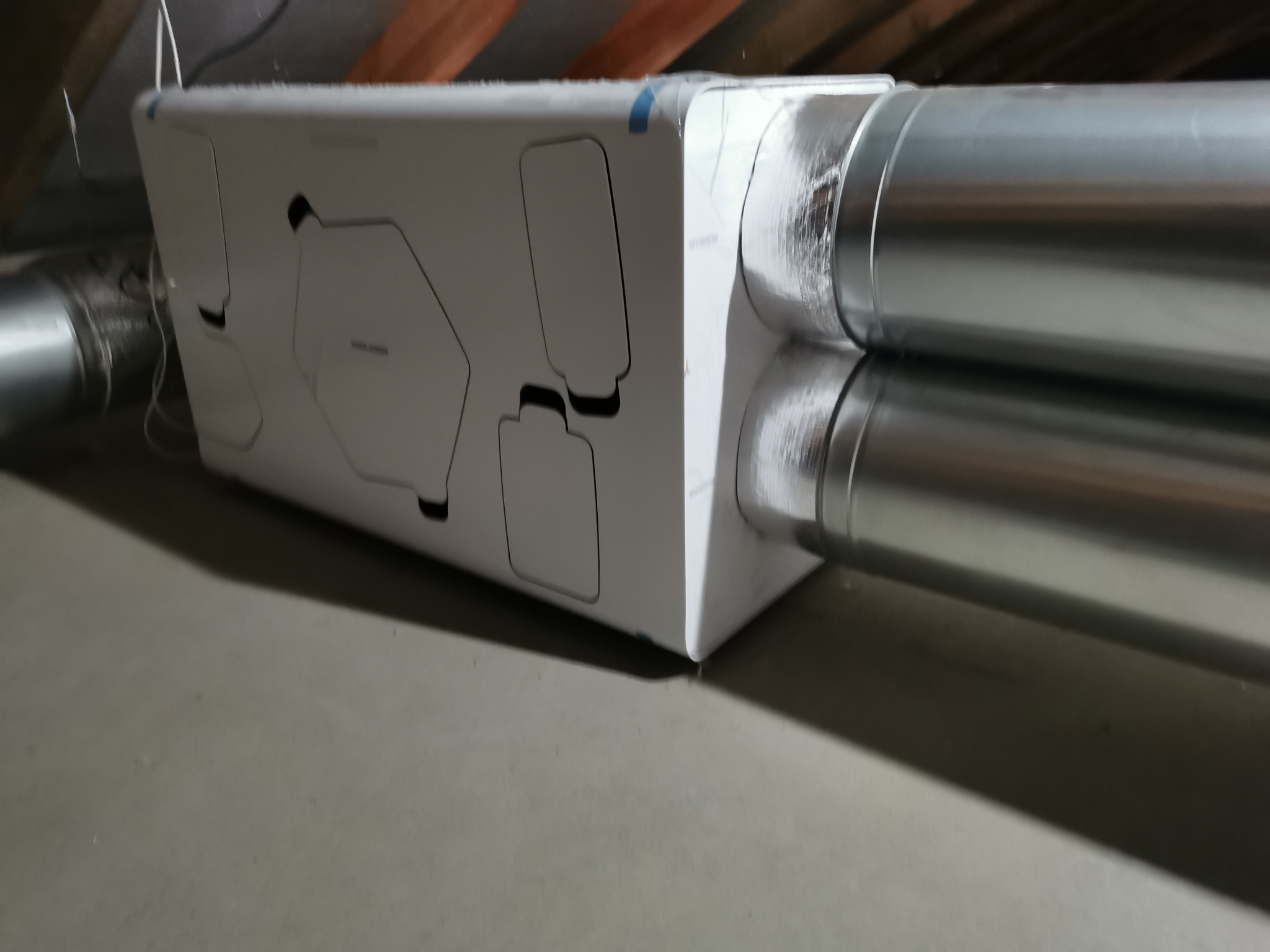 Centrala rekuperacyjna z podłączonymi przewodami wentylacyjnymi, zamontowana pod stropem. Widoczne detale obudowy i metaliczne rury wentylacyjne.