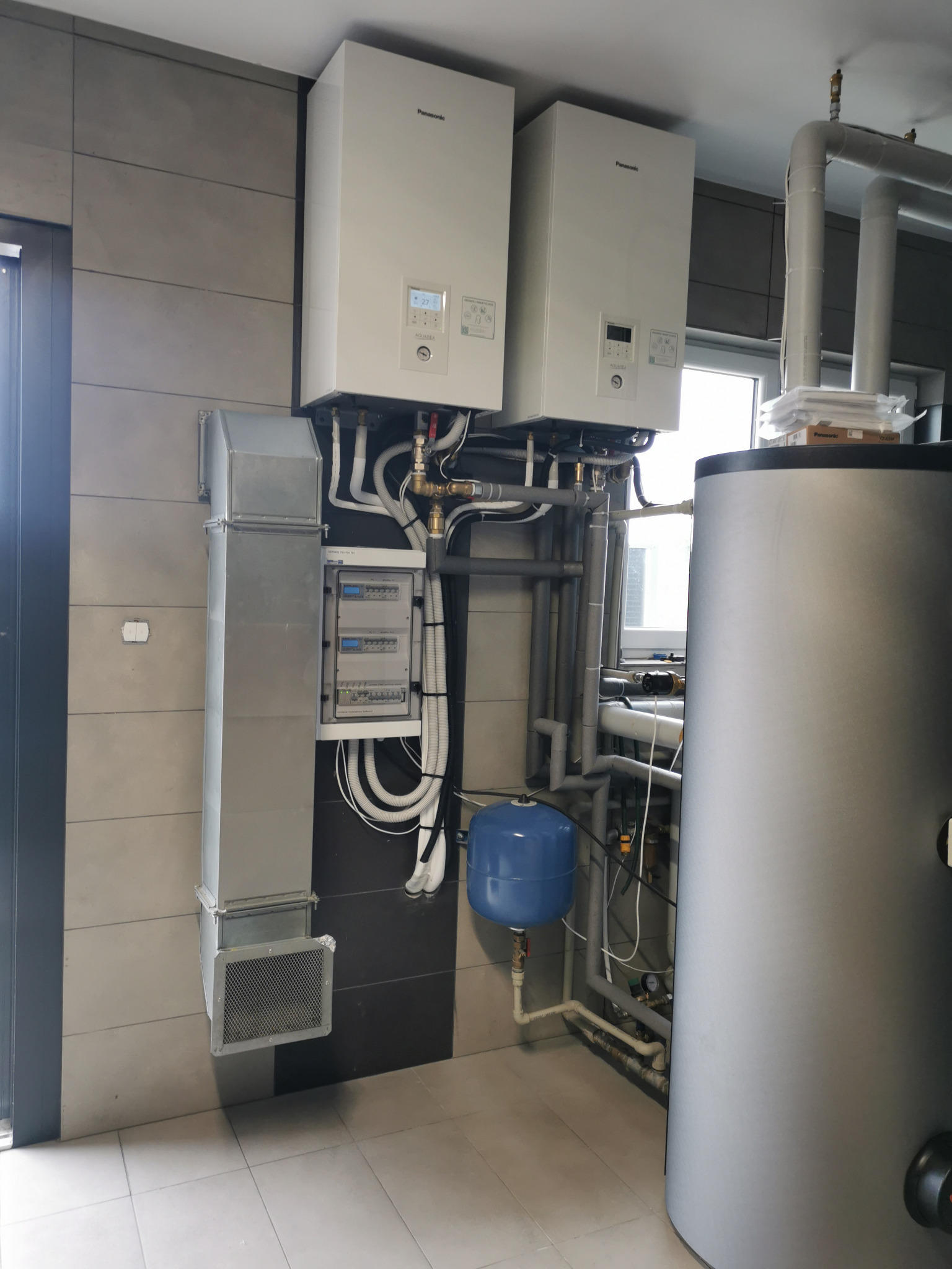 Dwie białe pompy ciepła Panasonic, szara instalacja rekuperacji z kanałami i niebieskim zbiornikiem, widoczne rury i zawory na tle szaro-białej ściany z płyt.